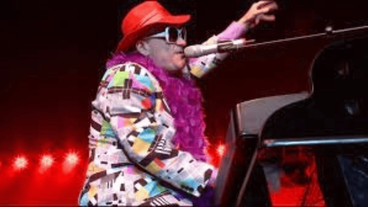 A tribute performer dressed as Elton John plays a grand piano. He is wearing a multicolored jacket, red cowboy hat, rhinestone sunglasses, and a purple feather boa.